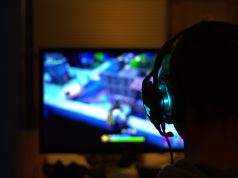 Gracz siedzący przed komputerem, na ekranie widoczna jest rozgrywka, gracz ma na uszach słuchawki
