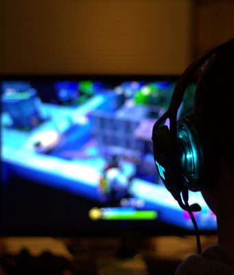 Gracz siedzący przed komputerem, na ekranie widoczna jest rozgrywka, gracz ma na uszach słuchawki